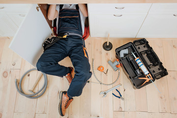 Man in uniform work in kitchen under sink. He repair water leaking. Hose and tools on floor. Opened...
