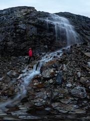man in red stands under a waterfall on a black rock in the mountains on a cloudy rainy day