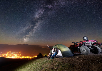 Young couple man and woman sitting together in tourist tent on the top of mountain with atv quad motorbike. Man pointing at beautiful night sky full of stars, Milky way, luminous town on background © anatoliy_gleb