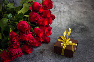 A bouquet of red roses next to a box with a gift, tied with a ribbon and sweets, a holiday greeting card, a festive background with flowers