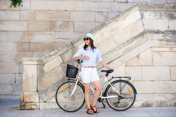 Fototapeta na wymiar Pretty smiling woman in sunglasses, white shorts, blouse and hat leaning on bicycle on white stone wall and stairs background on bright sunny day. Sightseeing and vacations concept.