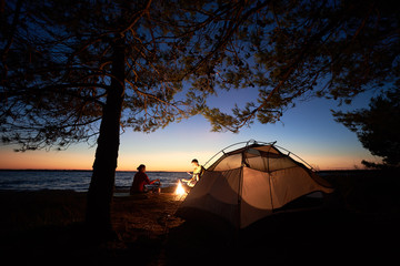 Camping on lake shore at night. Tourist tent under trees and young family, man and woman preparing food on campfire on dark blue evening sky and clear water background. Tourism and adventure concept.