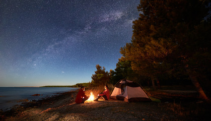 Night camping on shore. Young man and woman tourists sitting relaxed in front of tent at campfire...