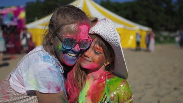 Teenage girl caring about her little sister put on a hat standing on the beach. Pretty children having fun at Holi festive covered in colorful dry paint. Happiness, summer journey, peaceful concept