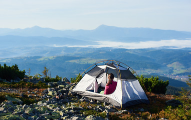 Camping on the top of mountain on bright summer morning. Back view of woman sitting in the entrance of tourist tent. On foggy mountains background. Tourism adventure holiday active lifestyle concept