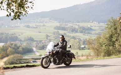 Long-haired bearded cool biker in sunglasses and black leather clothing riding cruiser powerful motorcycle along sunny asphalt road on bright summer day on background of rural misty landscape.