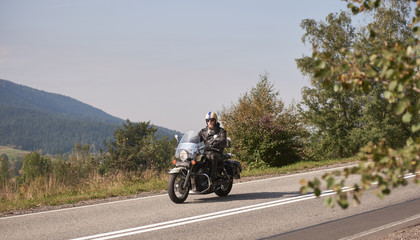 Fototapeta na wymiar Bearded biker in helmet, sunglasses and black leather clothing riding cruiser powerful motorcycle down sunny asphalt road on bright summer day on background of green woody hill.