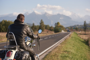 Back view of biker in black leather jacket riding cruiser motorcycle along road on blurred...