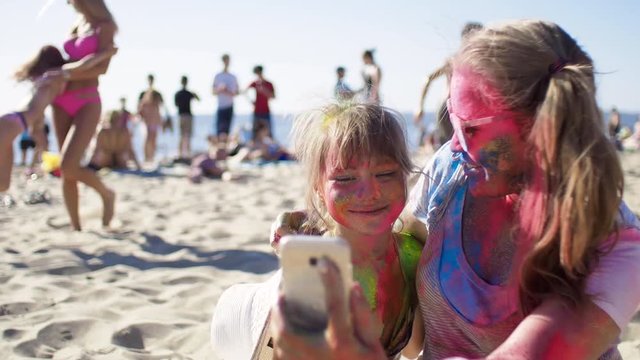 Pretty child and adult girl with colored faces doing photography to the smartphone at river coast, smile together and having fun. Colorful holi party on the vacation, sunny weather and people around