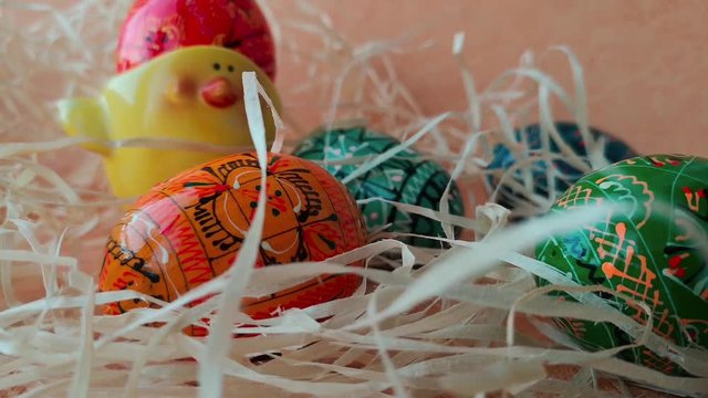 Happy Easter concept: multicolored painted Easter eggs and ceramic egg holder made in the shape of a chicken among decorative straw. The straw moves slightly in the wind.