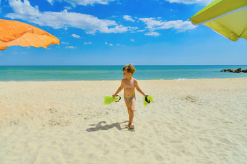 A little boy walks along the beach holding flippers in his hands.