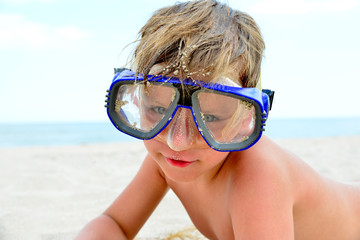 Fototapeta premium A cheerful and funny boy in flippers and a mask for scuba diving lies on the sand by the sea and sunbathes in the sun in a clear summer.