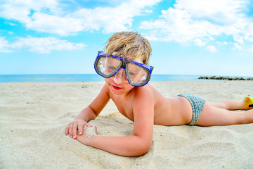 A cheerful and funny boy in flippers and a mask for scuba diving lies on the sand by the sea and sunbathes in the sun in a clear summer.