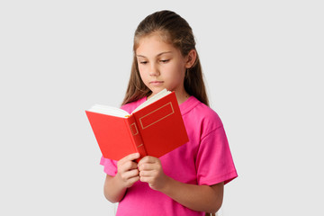 Smart little girl reading red book against a grey background. Lovely schoolgirl studying at home looking to the book attentively and learning material