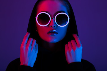 Portrait de mode jeune fille dans un pull noir avec une capuche et des lunettes de soleil rondes en néon rouge et bleu dans le studio