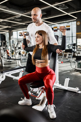 Fototapeta na wymiar Beautiful slender girl in stylish sports clothes does a dumbbell bench press up sitting on a bench while the athletic man insures her in the modern gym