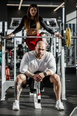 Fototapeta na wymiar Brutal athletic man sits on the sport bench and slender girl stands behind him next to the sport equipment in the modern gym