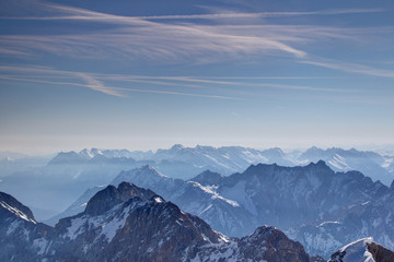 Jubilaumsgrat ridge in shadows and sunny hazy ridges of Wetterstein and Karwendel mountains under blue sky with clouds and contrails from Zugspitze Northern Limestone Alps Bayern Germany Tirol Austria