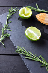 Fototapeta na wymiar Lime halves and wooden juicer on slate board. Lime and rosemary on black background. Fresh lime juice flows over the stone board. Healthy food