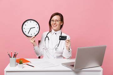 Female doctor sit at desk work on computer with medical document hold money in hospital isolated on pastel pink wall background. Woman in medical gown glasses stethoscope. Healthcare medicine concept.