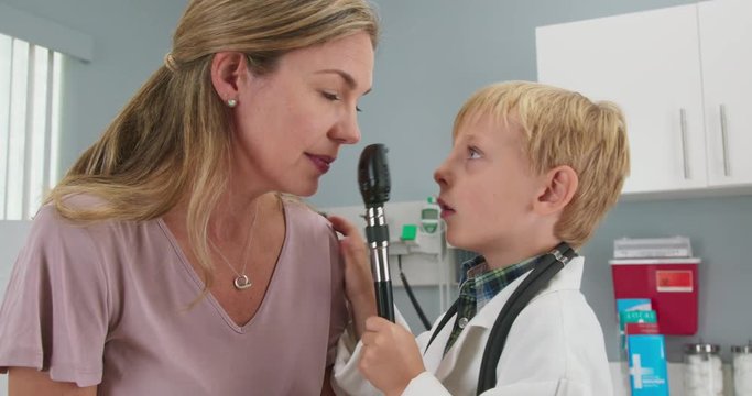 Cute little boy pretending to be a doctor with his mother while in pediatricians office. Child wearing oversized lab coat and stethoscope using ophthalmoscope. Slow motion 4k