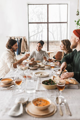 Group of beautiful international friends sitting at the table full of food talking to each other. Young colleagues dreamily spending time together on lunch with big window on background in cozy cafe