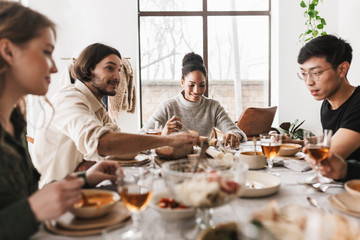 Group of attractive international friends sitting at the table full of food spending time in cozy...