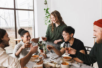 Pretty smiling woman with wavy hair holding glass of wine in hand standing near table full of food. Group of attractive international friends joyfully spending time together having lunch in cafe