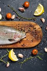 Obraz na płótnie Canvas Raw trout on a wooden chopping Board. Ingredients rosemary, lemon, tomatoes, garlic, salt, pepper. Black background, top view