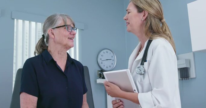 Senior patient laughing with her doctor in exam room. Female primary care physician talking to older Caucasian woman during regular check up. Slow motion 4k