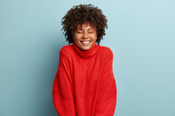 People and enjoyment concept. Pleased black woman expresses sincere emotions, wears casual red sweater, rejoices life, laughs out loudly, has fun and being amused, isolated over blue background