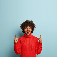 Photo of merry dark skinned woman shows direction upwards, wears red jumper, isolated over blue...