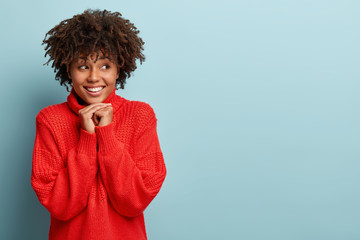 Excited joyful dark skinned girl has broad smile, shows white teeth, keeps hands together uтder chin, focused away, wears red sweater, enjoys good moment, isolated over blue wall with free space