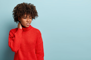 Sad dissatisfied young woman with dark skin, has regretful look, keeps hand on neck, wears red loose jumper, gazes down, dressed in red oversized jumper, isolated over light blue studio wall