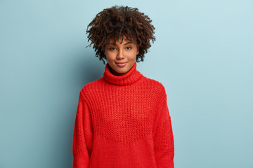 Headshot of beautiful young dark skinned woman with curly hair, dressed in red winter jumper, smiles pleasantly at camera, has no make up, expresses good feelings, isolated over blue background