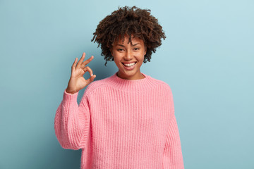 Headshot of positive dark skinned woman with crisp hair, makes okay gesture, wears loose sweater,...