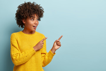 Merry curious black woman with crisp hair, wears yellow jumper, points with both fore fingers at empty space, demonstrates direction to nice place for entertainment, isolated over blue background
