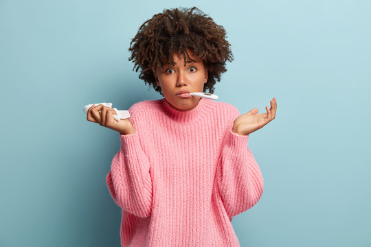 People, disease concept. Confused black woman with crisp hair, has high temperature, holds thermometer in mouth, tissue in hand, cant understand where she caught cold, isolated over blue background