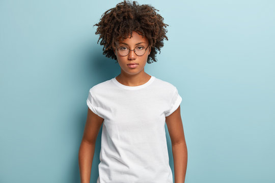Studio shot of serious thoughtful feminine girl with dark skin, crisp hair, looks directly at camera, wears spectacles and t shirt, thinks over problematic situation, isolated over blue background