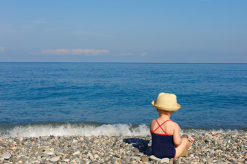 Fototapeta na wymiar Little girl in a bathing suit looking at sea, rear view, space for text. Charming baby sitting on the seashore, summer photo.