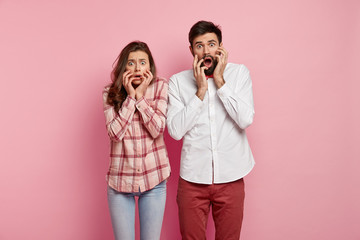Woman and man in awe look with scared facial expressions, keep hands on faces, have eyes full of...