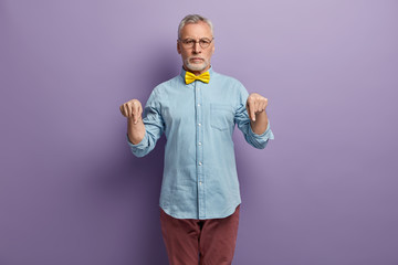 Image of self confident serious mature man with thick grey bristle, wears elegant shirt and trousers, points down, advertises something, isolated over purple studio background. Just look at floor