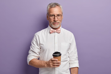 Shot of attractive elegant man with hot beverage, holds paper cup, wears elegant white shirt and pink bowtie, isolated over purple background, looks directly at camera. People, age, drink concept