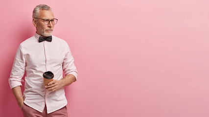 Waist up shot of attractive grey haired senior man with stubble, wears formal shirt, keeps hand in pocket, holds takeaway coffee, keeps gaze aside, isolated over rosy background with blank space