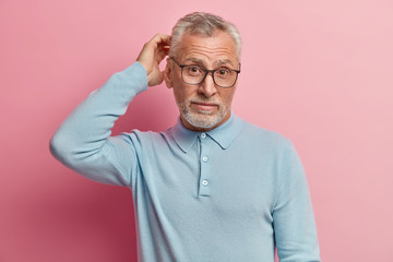 Shot of attractive bearded Caucasian man with thick grey stubble, scratches head in puzzlement,...