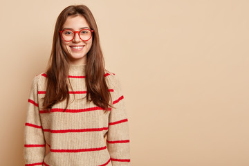 Studio shot of good looking woman with toothy smile, wears transparent glasses and sweater, being...
