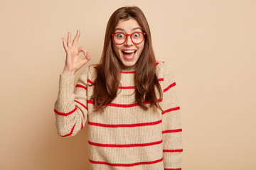 Isolated shot of happy delighted woman shows okay gesture, feels amazed, wears spectacles and...