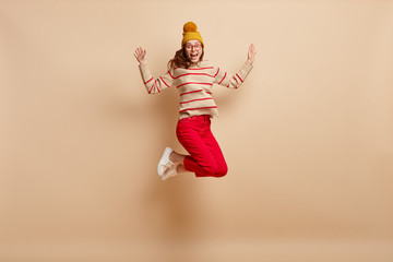 Emotional young woman with overjoyed expression, jumps high in air, wears yellow hat with pompon, striped sweater and red trousers, isolated over brown background, feels energetic and elated