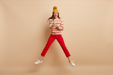 Shot of happy Caucasian woman points at camera, makes finger gun gesture, spreads legs, wears headgear, jumper, trousers ans sportshoes, jumps against beige background. Horizontal indoor image