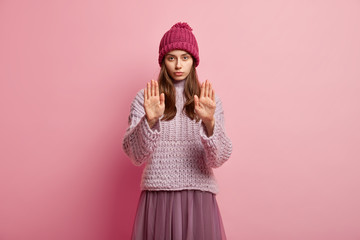 Image of serious young European woman demonstrates stop sign or prohibition symbol, stretches hands and shows palms, wears headgear, loose knitted sweater, skirt, models over rosy background